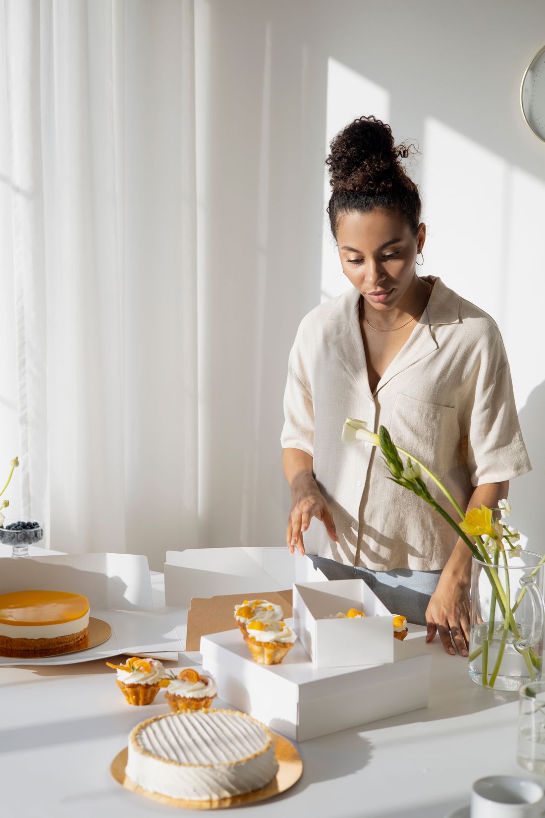 A baker boxing an order of cupcakes and a tart in a bright, sunlit kitchen